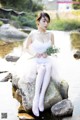 A woman in a white dress sitting on a rock by the water.